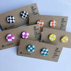 Puede incluir: Un conjunto de ocho pendientes con estampado de tablero de ajedrez en varios colores, incluyendo negro y blanco, rosa y blanco, azul verdoso y blanco, naranja y blanco, y amarillo y blanco. Los pendientes se muestran en tarjetas marrones con el texto "slab." y "@youslab" impreso en ellas.