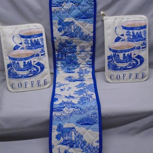 May include: A set of kitchen textiles featuring a blue and white toile pattern. The set includes two pot holders with a coffee cup design and a double oven mitt. The oven mitt and pot holders have a white background with blue trim.