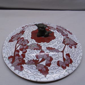 May include: A decorative, round lid with a white background and red floral design. The lid features a small, bronze-colored figurine on top. The floral pattern includes red and pink flowers and leaves. The lid's edge has a scalloped design.
