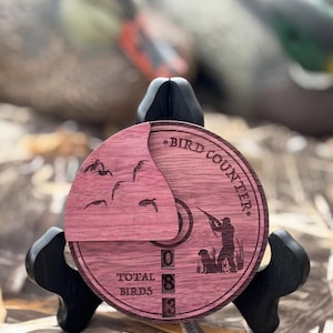 May include: A wooden bird counter with a pinkish-red stain, featuring a hunter silhouette, dog, and flying birds. The text "BIRD COUNTER" and "TOTAL BIRDS" are engraved. The counter shows the number 083.