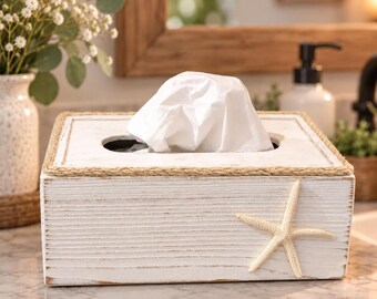 Könnte beinhalten: Ein rechteckiger Taschentuchbox-Halter aus weißem Holz mit Used-Look, verziert mit einer Kordel aus Naturjute am oberen Rand und einem cremefarbenen Seestern an der Vorderseite. Ein weißes Taschentuch ragt aus der mittigen Öffnung heraus.