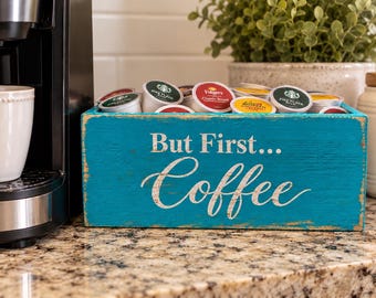 Könnte beinhalten: Eine rechteckige, türkisfarbene Holzaufbewahrungsbox mit abgenutzten Kanten und dem weißen Schriftzug "But First... Coffee" auf der Vorderseite. Die Box ist mit verschiedenen Kaffeekapseln gefüllt, darunter Marken wie Starbucks und Folgers.
