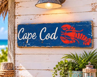 Könnte beinhalten: Ein rechteckiges Holzschild mit abgenutzter dunkelblauer Oberfläche, das den weißen Schriftzug Cape Cod und eine leuchtend rote Hummergrafik zeigt. Das Schild ist an einer weißen Wand aus horizontalen Brettern unter einer Metalllampe montiert.
