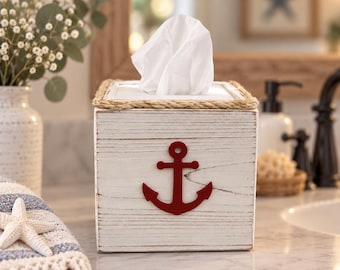 Könnte beinhalten: Ein quadratischer Taschentuchbox-Behälter aus Holz mit weißem Used-Look-Finish und einem roten Anker-Emblem auf der Vorderseite. Die Oberkante ist mit einer dicken, naturfarbenen Juteschnur verziert, und ein weißes Taschentuch ragt oben heraus.