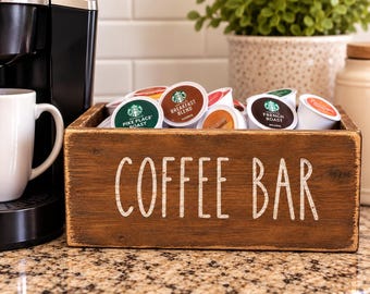 Könnte beinhalten: Eine rechteckige Holzkiste mit dem in Weiß aufgedruckten Text COFFEE BAR auf der Vorderseite. Die Kiste ist mit verschiedenen Starbucks-Kaffeekapseln gefüllt, darunter Pike Place Roast, Breakfast Blend und French Roast, und steht auf einer Granitarbeitsplatte.
