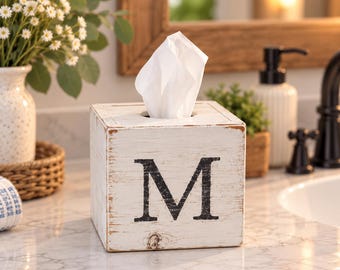 Könnte beinhalten: Ein quadratischer Taschentuchbox-Überzug aus Holz mit weißem Used-Look-Finish und einem großen, schwarzen Buchstaben M auf der Vorderseite. Die Box zeigt eine sichtbare Holzmaserung und abgenutzte Kanten, mit einem weißen Tuch in der oberen Öffnung.
