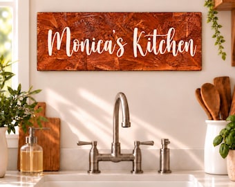 Könnte beinhalten: Ein rechteckiges Wandschild aus dunkelbrauner Grobspanplatte mit dem weißen Schriftzug "Monica's Kitchen" in Schreibschrift, montiert über einer Küchenarmatur aus Edelstahl und einem weißen Spülbecken.