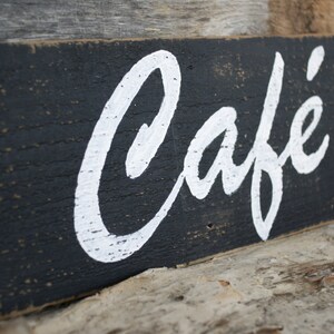 K&ouml;nnte beinhalten: Ein rechteckiges Holzschild mit abgenutztem schwarzem Finish und dem Wort Caf&eacute; in wei&szlig;er Schreibschrift. Die Holzmaserung ist durch die schwarze Farbe sichtbar und der wei&szlig;e Text hat ein strukturiertes, handgemaltes Aussehen.