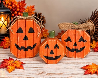 Könnte beinhalten: Drei orangefarbene Kürbisdekorationen aus Holz mit schwarz bemalten Jack-o'-Lantern-Gesichtern. Die Kürbisse haben grüne Stiele und sind mit Schleifen aus Bindfaden oder Jute verziert. Jeder Kürbis weist ein abgenutztes Finish auf.