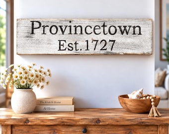 Könnte beinhalten: Ein rechteckiges Holzschild mit abgenutzter weißer Oberfläche und schwarzer Aufschrift Provincetown Est. 1727. Das Schild weist eine verwitterte Textur auf und ist horizontal an einer Wand über einem Holztisch montiert.