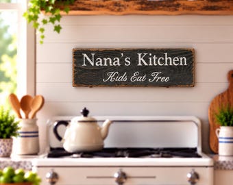 Könnte beinhalten: Ein rechteckiges Holzschild mit abgenutztem schwarzem Finish und weißer Aufschrift, die Nana's Kitchen und Kids Eat Free lautet. Das Schild weist eine sichtbare Holzmaserung auf und ist horizontal an einer weißen Wandverkleidung montiert.