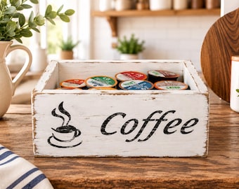 Könnte beinhalten: Eine rechteckige Aufbewahrungsbox aus weißem Holz mit Used-Look, die den schwarzen Schriftzug "Coffee" und eine Grafik einer dampfenden Tasse zeigt. Die Box ist mit verschiedenen bunten Kaffeekapseln gefüllt und dient als Küchenorganizer.