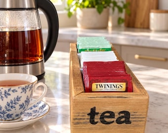 Könnte beinhalten: Eine rechteckige Teebeutel-Aufbewahrungsbox aus Holz mit dem Wort tea in schwarzer Schrift auf der Vorderseite. Die Box enthält Reihen von Teebeuteln in roter, weißer und grüner Verpackung, darunter ein Twinings-Teebeutel vorne.