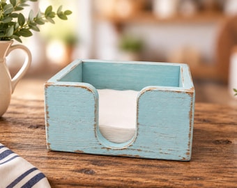 Könnte beinhalten: Ein quadratischer Serviettenhalter aus Holz mit einem abgenutzten hellblauen Finish und sichtbarer brauner Holzmaserung an den Kanten. Die Vorderseite verfügt über einen U-förmigen Ausschnitt für den einfachen Zugriff auf die weißen Servietten.