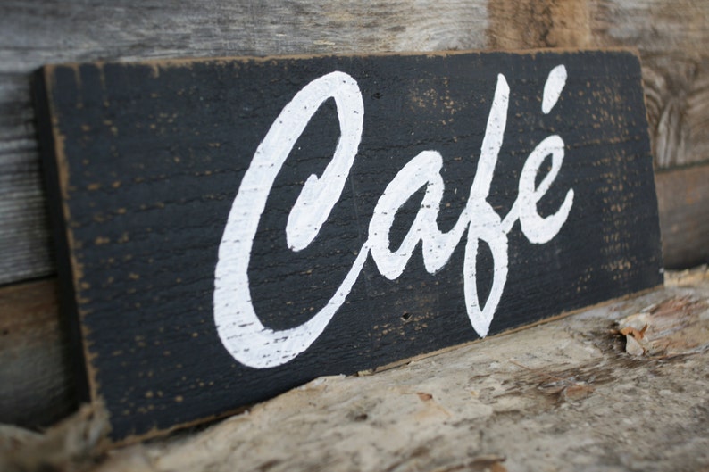 K&ouml;nnte beinhalten: Ein rechteckiges Holzschild mit abgenutztem schwarzem Finish und dem Wort Caf&eacute; in wei&szlig;er Schreibschrift. Die Holzoberfl&auml;che weist eine sichtbare Maserung und abgenutzte Kanten auf, was dem gesamten Objekt ein verwittertes Aussehen verleiht.