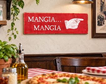 Könnte beinhalten: Ein rechteckiges rotes Holzschild mit abgenutzter Oberfläche, das den weißen Schriftzug MANGIA... MANGIA und eine weiße Grafik einer zeigenden Hand zeigt. Das Schild ist an einer strukturierten Wand über einem Tisch befestigt.