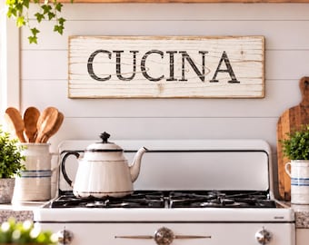 Könnte beinhalten: Ein rechteckiges Wand-Schild aus Holz mit weißem Used-Look-Finish und dem Wort CUCINA in schwarzen Großbuchstaben. Das Schild weist eine horizontale Holzmaserung auf und ist an einer weißen Paneelwand montiert.