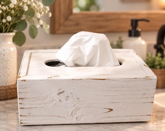 Könnte beinhalten: Ein quadratischer Taschentuchbox-Überzug aus Holz mit einem abgenutzten weißen Finish, das die natürliche braune Holzmaserung darunter freilegt. Die Box hat eine rechteckige Öffnung an der Oberseite, durch die ein weißes Papiertaschentuch ragt.