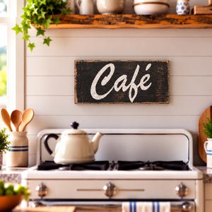 K&ouml;nnte beinhalten: Ein rechteckiges Wand-Schild aus Holz mit abgenutzter schwarzer Oberfl&auml;che und dem Wort Caf&eacute; in wei&szlig;er Schreibschrift. Das Schild weist eine verwitterte Textur auf und ist an einer wei&szlig;en Wand mit horizontalen Paneelen &uuml;ber einem Herd montiert.