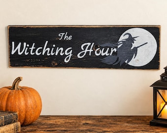 Könnte beinhalten: Ein rechteckiges schwarzes Holzschild mit abgenutzten Kanten, das den weißen Schriftzug The Witching Hour sowie die Silhouette einer Hexe auf einem Besen vor einem großen weißen Vollmond zeigt. Das Schild hat eine sichtbare Holzmaserung.