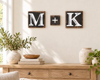 Könnte beinhalten: Drei quadratische Wandtafeln aus Holz mit abgenutzter schwarzer Oberfläche und weiß aufgemalten Buchstaben M und K sowie einem Pluszeichen in der Mitte. Die Schilder sind horizontal an einer Wand über einem Holzkonsolentisch angebracht.