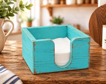 Könnte beinhalten: Ein quadratischer Serviettenhalter aus Holz mit abgenutzter türkisfarbener Lackierung. Der Halter verfügt über einen U-förmigen Ausschnitt an der Vorderseite und enthält einen Stapel weißer Papierservietten. Die Holzmaserung ist unter der Farbe sichtbar.