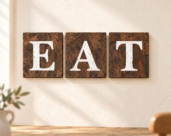 Könnte beinhalten: Drei quadratische Wandtafeln aus dunkelbraunen Grobspanplatten, die jeweils einen großen, abgenutzten weißen Großbuchstaben tragen. Zusammen bilden die Tafeln das Wort EAT in einer fetten Serifenschrift und sind horizontal an der Wand montiert.