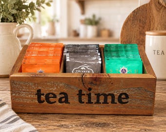 Könnte beinhalten: Eine rechteckige Tee-Aufbewahrungsbox aus Holz mit drei Fächern und dem aufgedruckten schwarzen Schriftzug "tea time" auf der Vorderseite. Die Box ist mit Reihen von orangefarbenen, grauen und grünen Teebeuteln gefüllt.