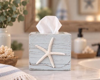 May include: A square light blue wooden tissue box cover featuring a textured, weathered finish and a prominent white starfish attached to the front center. The box holds a stack of white facial tissues extending from the top opening.