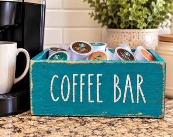 Könnte beinhalten: Eine rechteckige Aufbewahrungsbox aus blaugrünem Holz mit dem in Weiß aufgedruckten Text COFFEE BAR auf der Vorderseite. Die Box ist mit verschiedenen Kaffeekapseln in unterschiedlichen Farben gefüllt, darunter Starbucks Breakfast Blend.