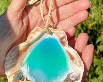 Peut inclure: Ornement en coquillage avec un motif en résine turquoise et blanche évoquant une scène de plage. La coquille est ornée d'une étoile de mer, de petits coquillages et d'éléments sableux. Une boucle en raphia naturelle est attachée.