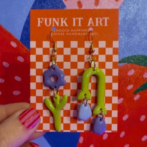 May include: A pair of handmade earrings with gold-colored hooks. One earring features a purple flower and a green, three-pronged shape. The other earring has a green arch and a blue teardrop shape. The earrings are displayed on a card that says "FUNK IT ART".