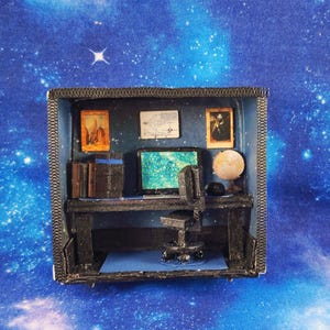 May include: A miniature diorama featuring a dark-toned office setup inside a box. The scene includes a black desk, a computer monitor with a green screen, a small office chair, a globe, and three framed pictures hanging on the back wall.