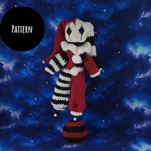 May include: A crocheted jester doll with a black and white striped body, red and white hat, and a black and white ball. The doll is standing on a blue background with white stars.