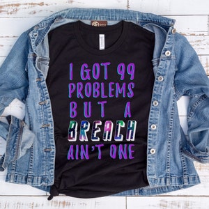 Op de afbeelding: Zwart t-shirt met de tekst "I GOT 99 PROBLEMS BUT A BREACH AIN'T ONE" in paars, blauw en groen. Het shirt wordt gepresenteerd op een wit houten oppervlak met een spijkerjasje.