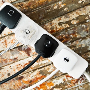 Puede incluir: Una regleta blanca con cuatro enchufes. Los enchufes son negros, blancos y transparentes. La regleta está sobre una superficie de madera.