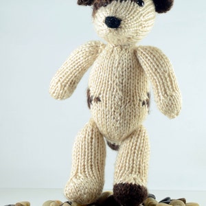 May include: A knitted beige dog toy with brown accents, standing on a bed of small, smooth stones.