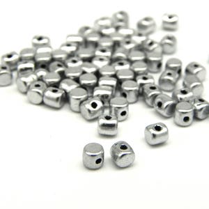 Könnte beinhalten: Ein Haufen silberner Metallperlen mit einem Loch in der Mitte jeder Perle. Die Perlen sind auf einer weißen Oberfläche verstreut.