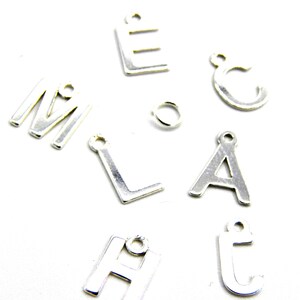 Op de afbeelding: Zilveren letterbedels in de alfabetletters E, M, L, A, H, C en T. De bedels zijn op een witte achtergrond.