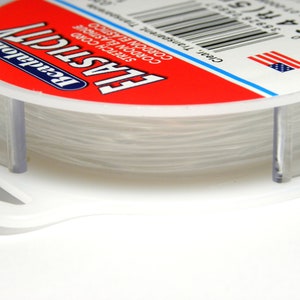 May include: A spool of clear elastic cord, labeled "Beadalon Elasticity" with the text "Clear, Transparent" and "16.4M/5" printed on the label. The spool is made of clear plastic and has a white label with red and blue accents.
