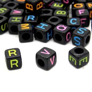 May include: Black square beads with colourful letters. The beads have a hole through the centre and are scattered on a white surface. The letters are in various colours, including green, pink, orange, and blue.