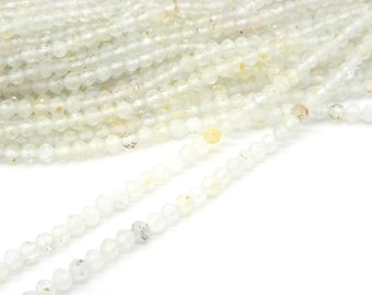 May include: Strands of small, faceted, off-white beads. The beads have a subtle sparkle and a slightly irregular shape, suggesting a natural material. The beads are strung together, creating a flowing, textured appearance. The background is white.