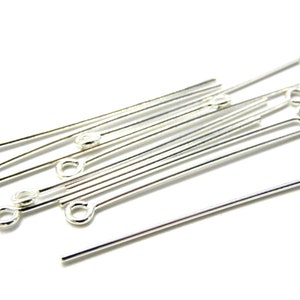 May include: A group of silver head pins with a loop at one end. The pins are arranged in a random pattern on a white surface.