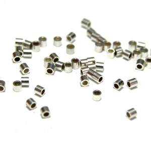 May include: Scattered assortment of small, silver-colored metal tube beads. Each bead is cylindrical with a hole running through the center. The beads are scattered on a white surface, with varying orientations.