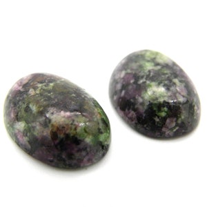 Könnte beinhalten: Zwei ovale Edelsteine mit polierter Oberfläche. Die Steine zeigen eine Mischung aus Farben, darunter Dunkelgrün, Lila und Schwarz, mit einem gesprenkelten Muster. Die Edelsteine sind vor einem weißen Hintergrund platziert.