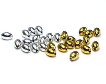 May include: A collection of silver and gold-colored oval beads. The beads are smooth and shiny, with a small hole at each end. They are scattered on a white surface, suitable for jewelry making or crafts.