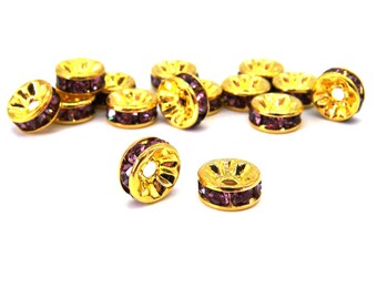 May include: A collection of gold-colored spacer beads with a ring of purple rhinestones. The beads have a decorative, openwork design on the sides. The beads are scattered on a white background.