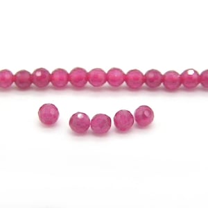 Peut inclure: Un rang de perles roses vives à facettes, avec plusieurs perles en vrac dispersées en dessous. Les perles sont rondes et ont une surface à facettes multiples, reflétant la lumière. Les perles sont enfilées sur un cordon fin et transparent, sur fond blanc.