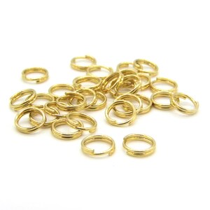Puede incluir: Un conjunto de pequeños anillos partidos dorados, dispersos sobre una superficie blanca. Los anillos son de doble bucle, diseñados para conectar componentes de joyería u otros objetos pequeños. La imagen muestra una vista de cerca de los anillos.