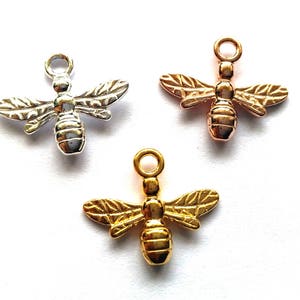 Könnte beinhalten: Drei Bienen-Anhänger in verschiedenen Metalloberflächen: Silber, Gold und Roségold. Jeder Anhänger zeigt ein detailliertes Bienendesign mit Flügeln und einem strukturierten Körper.